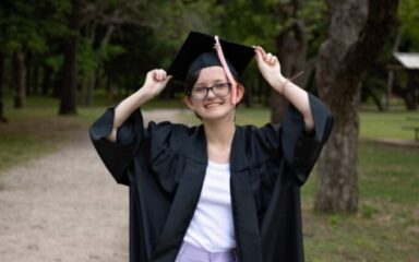 Una graduada sonriente se encuentra al aire libre en un camino de grava, sujetando los lados de su birrete de graduación con ambas manos. Lleva toga y birrete negros sobre un top blanco y pantalones lavanda. Los árboles y la hierba rodean el camino, creando una escena tranquila y festiva.