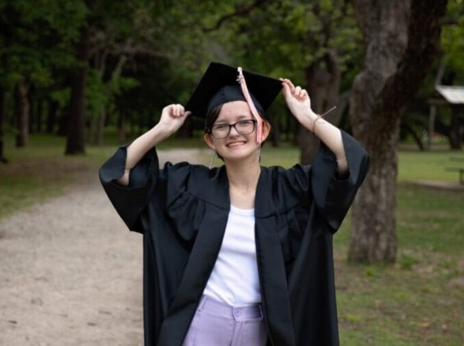 Una graduada sonriente se encuentra al aire libre en un camino de grava, sujetando los lados de su birrete de graduación con ambas manos. Lleva toga y birrete negros sobre un top blanco y pantalones lavanda. Los árboles y la hierba rodean el camino, creando una escena tranquila y festiva.