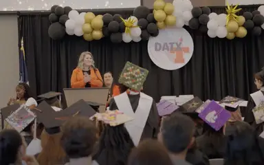 Foto de la graduación de DATX. Detrás de los estudiantes con un administrador en el podio hablando.