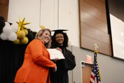 Un graduado con toga y birrete negro sonríe y recibe un diploma de manos de una mujer con chaqueta naranja.