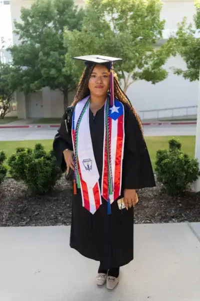 Una graduada luce orgullosa su toga y birrete negros, adornados con un fajín de la bandera de Puerto Rico y cordones de honor.