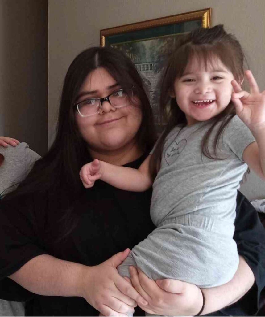 Persona con camisa negra sosteniendo a un niño con un traje gris con un diseño de corazón, en interiores con una foto enmarcada en la pared.